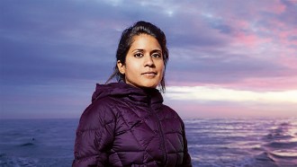 A headshot of Asha de Vos, a marine biologist from Sri Lanka. She is staring right into the camera and her black hair is tied to the back. She is wearing a purple puffy jacket and standing in front of the ocean.