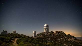 A line of four telescopes on a mountain ridge under a starry sky